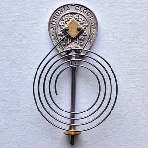 May include: Ansonia Clock Co. clock part. The silver-toned metal piece features the company name and a decorative design. A gold-colored square is centered on the top. A dark metal spiral and rod extend downward.