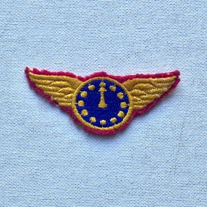 May include: Embroidered patch featuring a clock design with wings. The clock face is blue with a gold-colored clock hand and hour markers. The wings are gold with red trim, creating a vintage aesthetic. The patch is likely for a uniform or apparel.