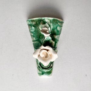 May include: A ceramic wall vase with a green and white marbled design. A white ceramic rose and bud are attached to the front of the vase. The vase is shaped like a small, open-topped cone.