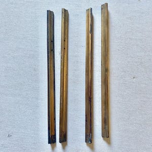 May include: Four antique brass picture frame pieces. The rectangular strips have varying shades of gold and brown, with visible wear and tear. The pieces are arranged vertically against a white background, suggesting a vintage or repurposed theme.