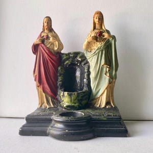 May include: A religious figurine featuring two standing figures flanking a central grotto-like structure on a black base. The figure on the left wears a red robe, and the figure on the right wears a pale green robe. Both figures hold a red heart.