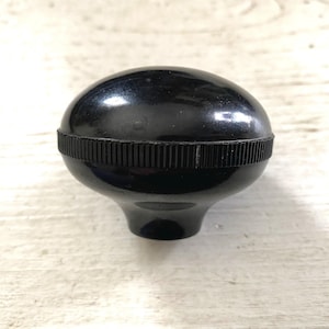 May include: A glossy black knob with a rounded top and a ribbed band around the middle. The knob has a smooth, reflective surface and a flared base. The object is likely a furniture or appliance component.