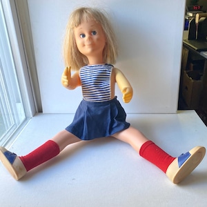 May include: A vintage doll with blonde hair, blue eyes, and a smiling expression. The doll wears a blue and white striped top, a navy blue skirt, red socks, and blue and beige shoes. The doll's legs are spread wide.