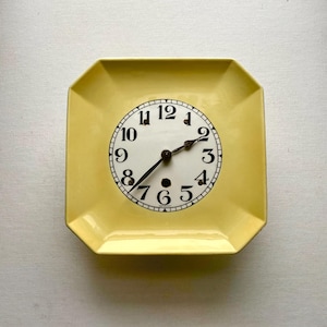 May include: A square, yellow clock with a white face and black numerals. The clock hands are black and point to 2:09. The clock is set in a square, yellow frame.
