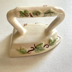 May include: Cream-colored ceramic iron with a curved handle and a flat base. The iron is decorated with green leaves and brown vines. A decorative piece for the home.