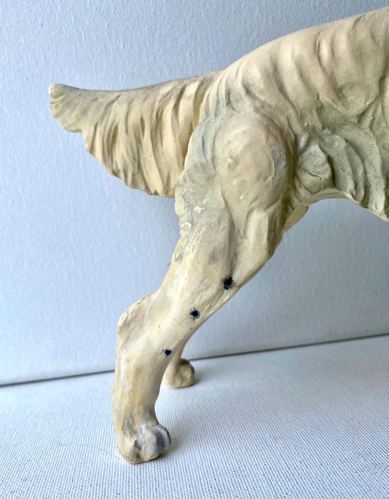May include: A cream-colored, vintage-style dog sculpture, showcasing the hindquarters and a portion of the tail. The sculpture has a textured surface with visible details and dark spots. The dog's leg is slightly bent, suggesting movement.