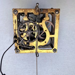 May include: Close-up of a vintage clock movement with brass frame and intricate gears. The gears are made of metal and are various sizes. A black wire and a silver pendulum are attached. The text "MADE IN GERMANY" is visible.