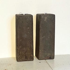 May include: Two rectangular, dark brown, weathered blocks. Each block is tall and narrow, with a slightly rough texture. The blocks are placed side by side on a light-colored surface, suggesting a rustic or industrial aesthetic.