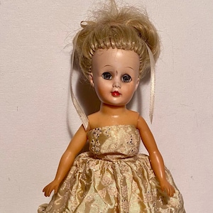 May include: Vintage doll with blonde hair styled in a bun, wearing a gold-colored dress with a floral pattern. The doll has painted facial features, including dark eyes and red lips. Two ribbons are attached to the doll's hair.
