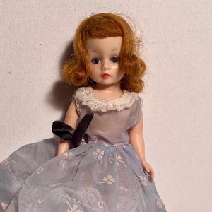 May include: A vintage doll with red hair styled in curls. The doll wears a light blue dress with a white lace collar and a black bow. The dress has a floral pattern on the skirt. The doll has a pale complexion and is in good condition.
