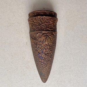May include: A bronze-colored, cone-shaped wall pocket with embossed grape and leaf designs. The top has a decorative band, and the bottom tapers to a point. This vintage-style piece is suitable for home decor.