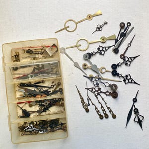 May include: A collection of vintage clock hands in various shapes and sizes, including gold, silver, and black. Some hands are ornate with decorative designs, while others are simple and streamlined. A clear plastic organizer holds additional clock hands.