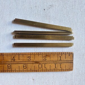May include: Several small, rectangular brass pieces are arranged near a wooden ruler. The ruler is marked with inch measurements. The brass pieces appear to be approximately 1/2 inch wide and vary in length.