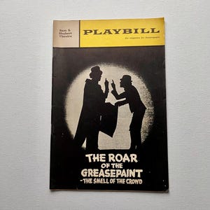 May include: A vintage Playbill magazine for "The Roar of the Greasepaint - The Smell of the Crowd." The cover features a black and white silhouette of two figures in hats, pointing at each other, against a white circle. The magazine has a yellow banner.