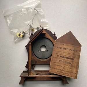 Puede incluir: Un reloj de madera antiguo, parcialmente desmontado, con un acabado oscuro. La esfera del reloj es visible, junto con una etiqueta de papel suelta y una peque&ntilde;a bolsa de piezas del reloj. La etiqueta dice "ONE DAY PANSY. F. KROEBER. CLOCKS, NEW YORK."