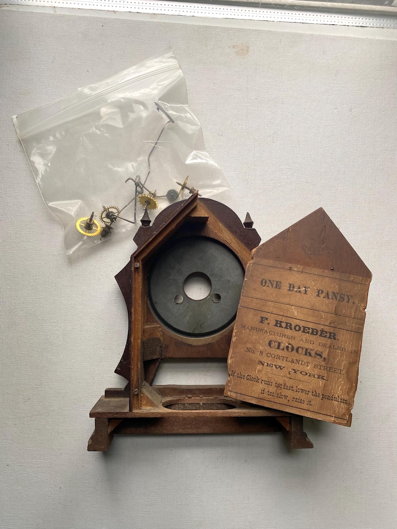 Puede incluir: Un reloj de madera antiguo, parcialmente desmontado, con un acabado oscuro. La esfera del reloj es visible, junto con una etiqueta de papel suelta y una peque&ntilde;a bolsa de piezas del reloj. La etiqueta dice "ONE DAY PANSY. F. KROEBER. CLOCKS, NEW YORK."
