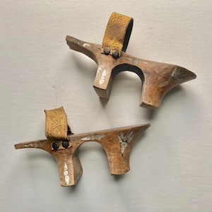 May include: A pair of wooden platform sandals featuring high arched soles with inlaid mother-of-pearl floral and geometric patterns. Each sandal has a textured mustard yellow fabric strap secured with two small dark metal studs on the wooden base.