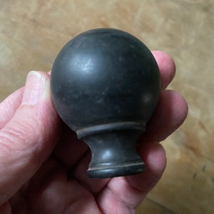May include: A dark gray, spherical finial with a decorative base. The finial has a smooth, matte finish and is likely made of wood or a similar material. The base has a classic, turned design.