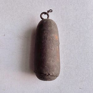 May include: A weathered, cylindrical metal object with a loop at the top. The dark brown piece has a slightly textured surface and a rounded base. The loop is made of a thinner, darker metal.