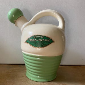 May include: A cream and light green ceramic decanter with a matching spherical stopper. The body features a label reading Mohawk Green Creme de Menthe, 80 proof, artificially colored, 25/32 of a quart, made and bottled by Mohawk Liqueur Corporation.