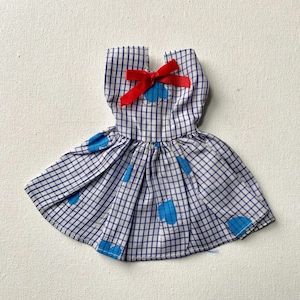May include: A doll-sized dress with a blue and white checkered pattern. The dress features a red bow at the neckline and blue patches on the skirt. The dress is made of fabric.