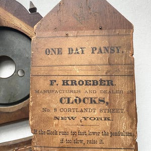 Puede incluir: Reloj de madera antiguo con una etiqueta de papel. La etiqueta dice "ONE DAY PANSY" y "F. KROEBER, FABRICANTE Y DISTRIBUIDOR DE CLOCKS, No. 8 CORTLANDT STREET, NEW YORK." Las instrucciones est&aacute;n impresas debajo. El reloj tiene un marco de madera oscura.