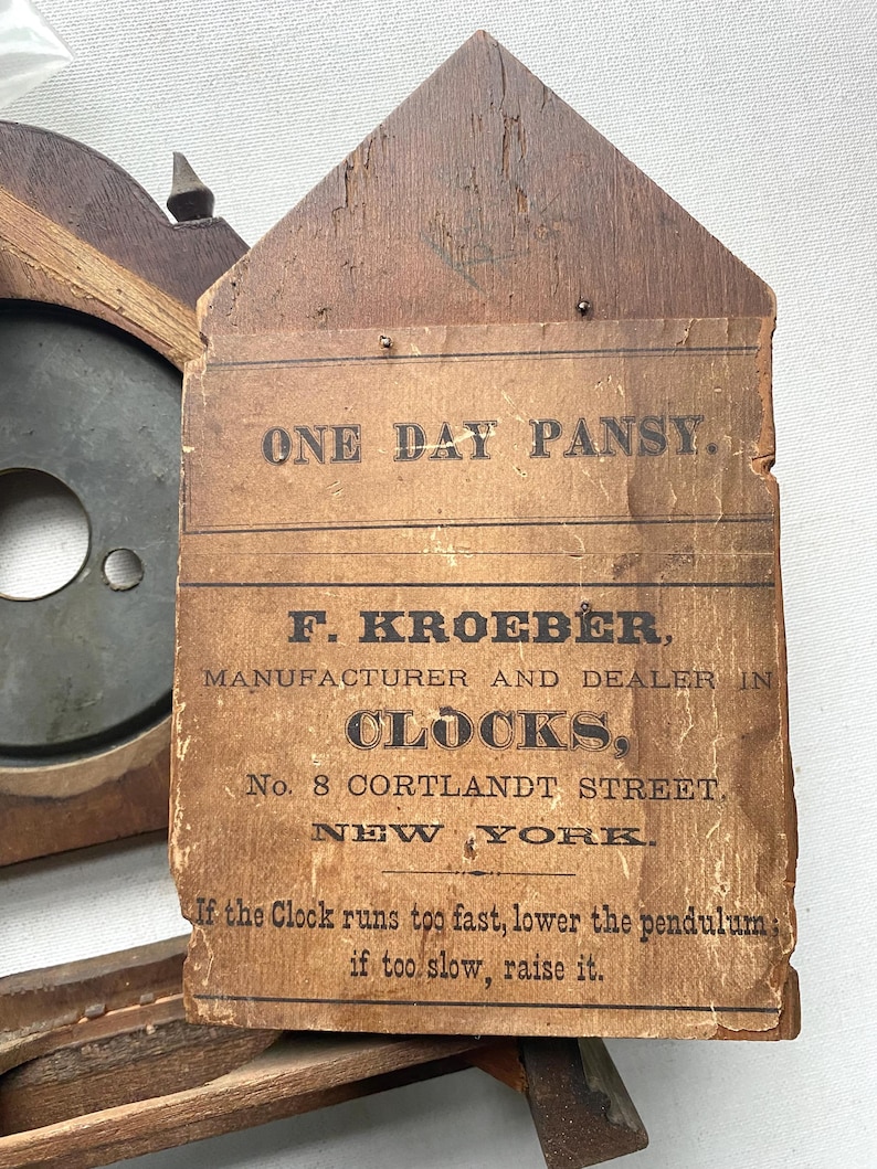 Puede incluir: Reloj de madera antiguo con una etiqueta de papel. La etiqueta dice "ONE DAY PANSY" y "F. KROEBER, FABRICANTE Y DISTRIBUIDOR DE CLOCKS, No. 8 CORTLANDT STREET, NEW YORK." Las instrucciones est&aacute;n impresas debajo. El reloj tiene un marco de madera oscura.