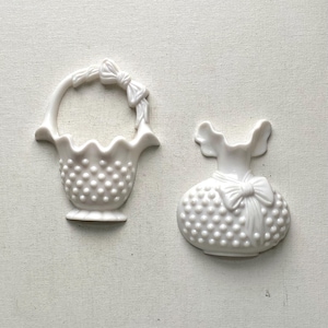 May include: Two white ceramic decorative pieces. One is a basket with a handle and bow detail. The other is a vase-shaped piece with a bow. Both have a textured, dotted surface.
