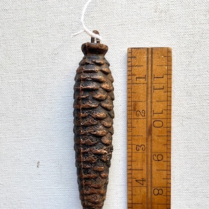May include: A bronze-colored pinecone ornament, approximately 4 inches long, with a textured surface. A wooden ruler is next to the ornament. A tag with the text "P6 MARKED 18" is attached.