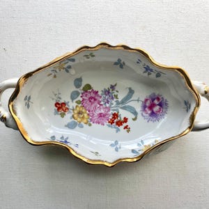 May include: Oval-shaped, white porcelain dish with a scalloped gold rim and two handles. The dish is decorated with a floral pattern in shades of pink, purple, yellow, and red. A decorative piece for serving.