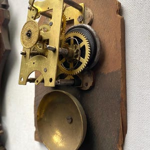 Puede incluir: Primer plano de un movimiento de reloj antiguo. El mecanismo presenta engranajes de lat&oacute;n, un resorte en espiral y una campana. El movimiento est&aacute; montado sobre una base de madera marr&oacute;n desgastada. El intrincado dise&ntilde;o del reloj muestra su edad y artesan&iacute;a.