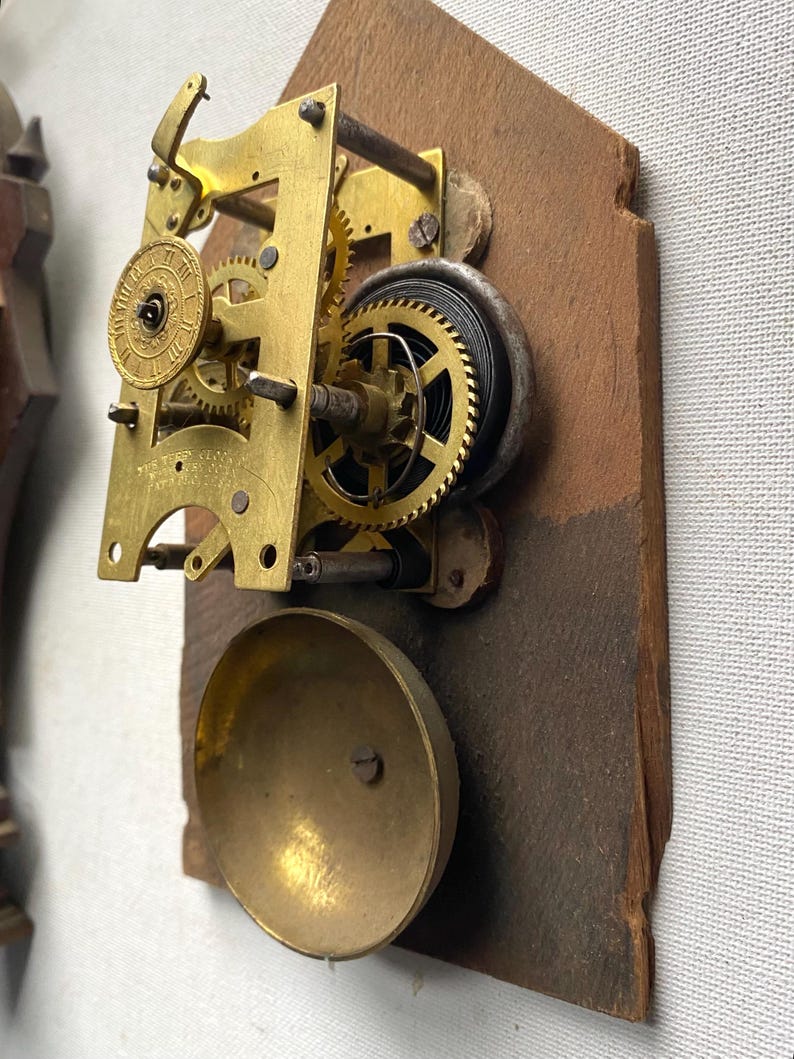 Puede incluir: Primer plano de un movimiento de reloj antiguo. El mecanismo presenta engranajes de lat&oacute;n, un resorte en espiral y una campana. El movimiento est&aacute; montado sobre una base de madera marr&oacute;n desgastada. El intrincado dise&ntilde;o del reloj muestra su edad y artesan&iacute;a.