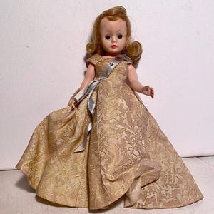 May include: A vintage doll in a gold-colored gown with a floral pattern. The doll has blonde hair styled in curls and is wearing a sash with a red jewel. The dress has a full skirt and short sleeves.
