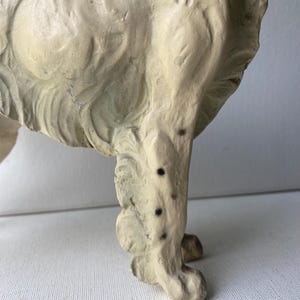 May include: A vintage, off-white dog figurine, possibly ceramic or plaster, with visible texture and wear. The dog's leg is in focus, showing details of the paw and the textured surface. Black spots are visible on the leg.