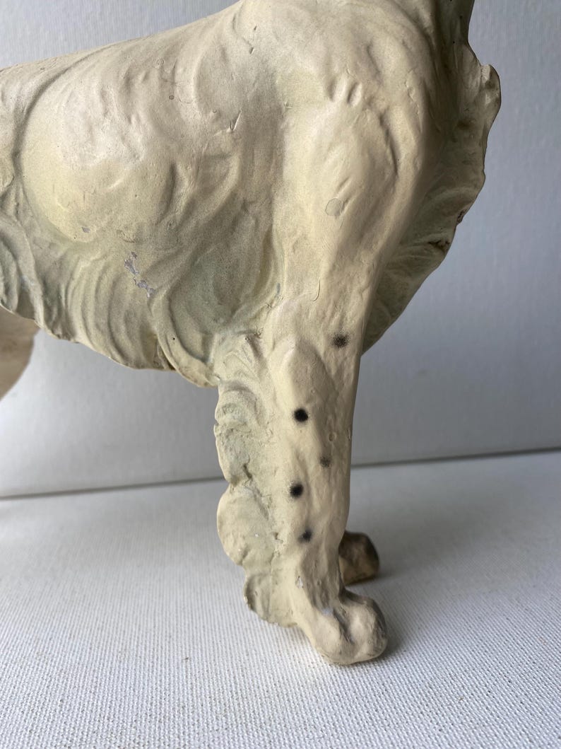 May include: A vintage, off-white dog figurine, possibly ceramic or plaster, with visible texture and wear. The dog's leg is in focus, showing details of the paw and the textured surface. Black spots are visible on the leg.