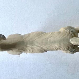 May include: A cream-colored dog figurine, lying down with its head raised. The dog's nose and ears are painted dark blue. The figurine has a long, pointed tail and is resting on a white surface.