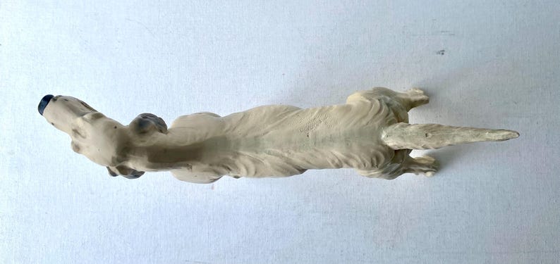 May include: A cream-colored dog figurine, lying down with its head raised. The dog's nose and ears are painted dark blue. The figurine has a long, pointed tail and is resting on a white surface.