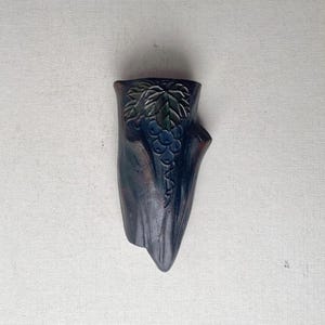 May include: A dark, hand-carved wooden wall pocket with a grape vine design. The pocket is shaped like a tree trunk, with a carved bunch of grapes and leaves. The colors are dark blue, green, and brown.