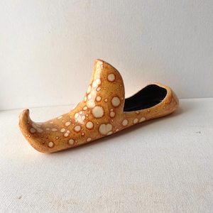 May include: A ceramic shoe-shaped vase with a speckled pattern in shades of tan and cream. The interior is black. The vase has a curved toe and a tall, curved heel.