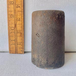 May include: A cylindrical, dark brown, textured metal weight positioned next to a wooden ruler. The ruler shows the object stands at a height of approximately 3.5 inches. The metal surface appears weathered with a rough, uneven, and oxidized finish.
