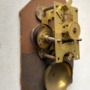 Puede incluir: Primer plano de un mecanismo de reloj antiguo. Los engranajes y componentes de lat&oacute;n est&aacute;n montados sobre una base de madera. El mecanismo del reloj incluye una campana, engranajes y un volante. El mecanismo es un bello ejemplo de artesan&iacute;a.