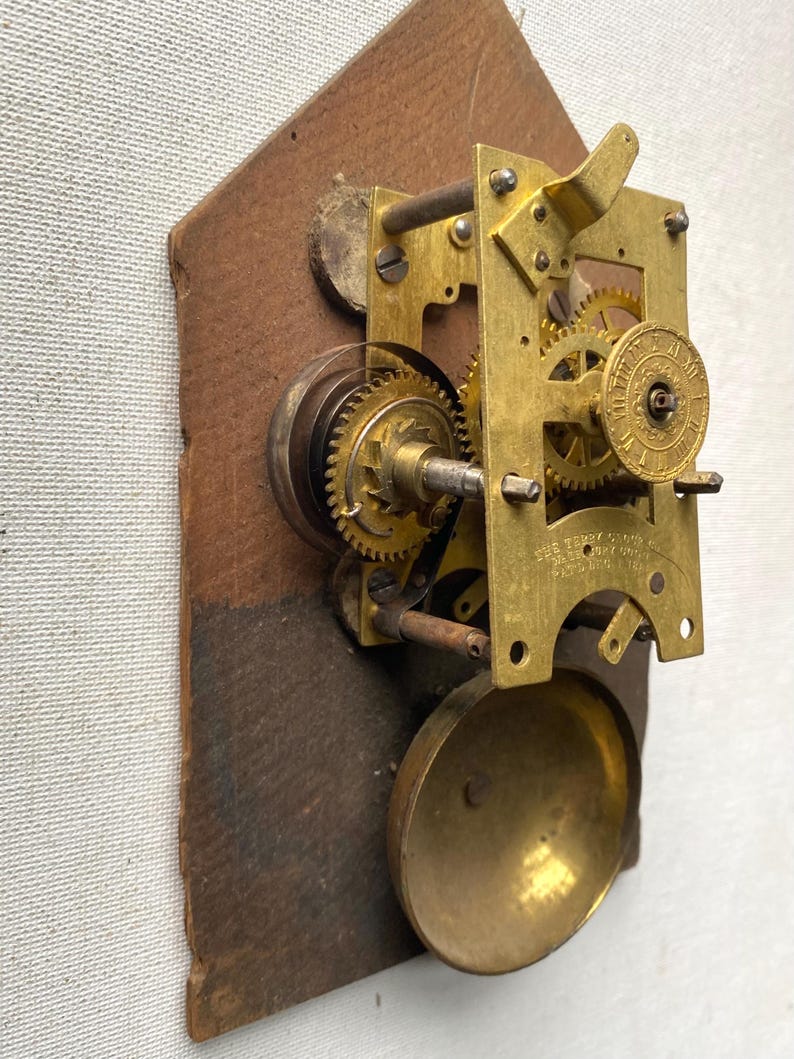 Puede incluir: Primer plano de un mecanismo de reloj antiguo. Los engranajes y componentes de lat&oacute;n est&aacute;n montados sobre una base de madera. El mecanismo del reloj incluye una campana, engranajes y un volante. El mecanismo es un bello ejemplo de artesan&iacute;a.