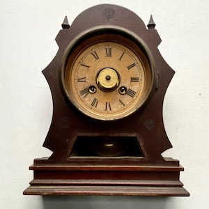 Puede incluir: Reloj de madera antiguo con acabado marr&oacute;n oscuro. La esfera del reloj es de color beige claro con n&uacute;meros romanos y manecillas negras. El reloj tiene un p&eacute;ndulo y est&aacute; sobre una base rectangular.