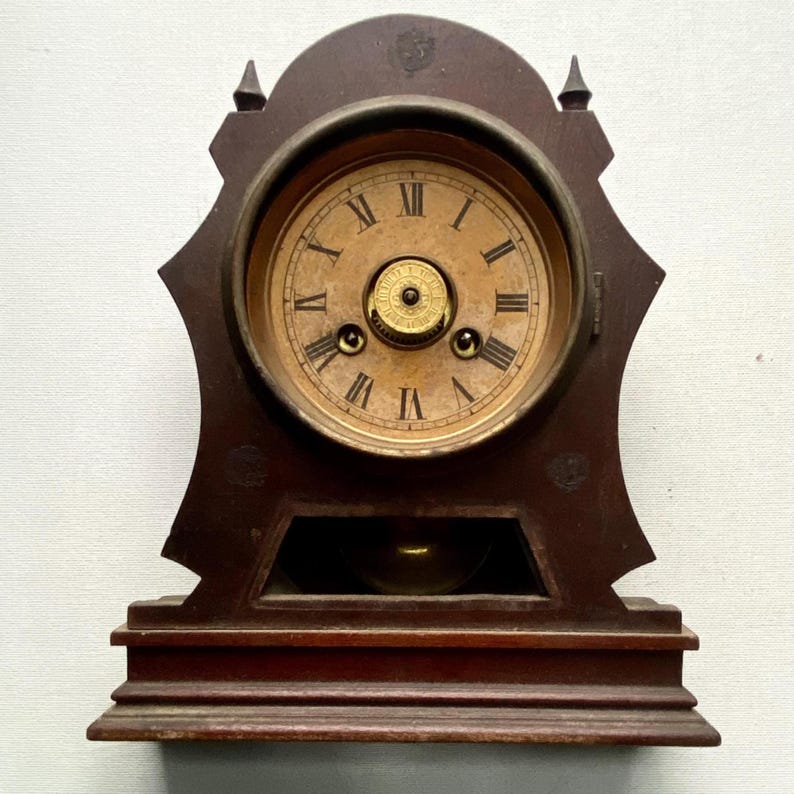 Puede incluir: Reloj de madera antiguo con acabado marr&oacute;n oscuro. La esfera del reloj es de color beige claro con n&uacute;meros romanos y manecillas negras. El reloj tiene un p&eacute;ndulo y est&aacute; sobre una base rectangular.