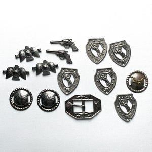 May include: A collection of silver-toned metal embellishments, including shields, guns, birds, and a buckle. The embellishments have various designs, including a bull's head, a bird, and a gun.