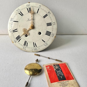 May include: A round white Zenith clock face with black Roman numerals and Arabic minute markers, featuring ornate gold hands. Included are a brass pendulum, a metal rod, and a rectangular paper manual with red and black graphics.