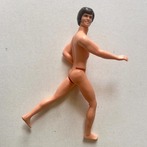 May include: A vintage plastic doll of a man in a running pose. The doll has short dark hair and is nude, with a tan skin tone. The doll is in a running pose with one leg extended forward.