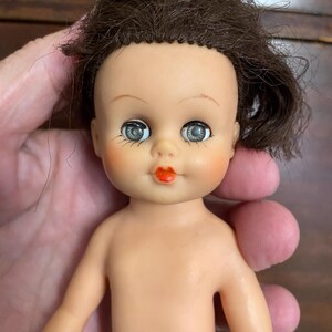 May include: A doll with a light skin tone, brown hair, and blue eyes. The doll features painted facial details including arched eyebrows, long eyelashes, and bright orange lips. The doll has a smooth, molded plastic body with a neutral expression.