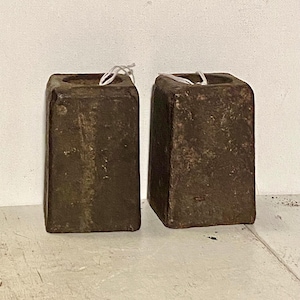 May include: Two weathered, rectangular, dark brown blocks with a square hole at the top. Each block has a white string tied around the top. The blocks are on a white surface.