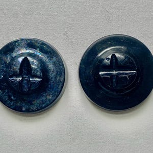 May include: Two circular buttons with a dark blue finish. The left button features a mottled, textured surface, while the right button has a smooth, glossy appearance. Both buttons have a central cross-shaped indentation for attachment.